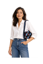 A woman wearing a white button-down shirt and blue jeans, carrying a small black shoulder bag with a colorful geometric pattern and a dark blue strap, set against a plain white studio background.