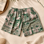 A pair of women's shorts with a green tropical palm tree and geometric pattern on a light pink background, styled in a flat-lay with a woven mat, book, slippers, and a knit blanket.