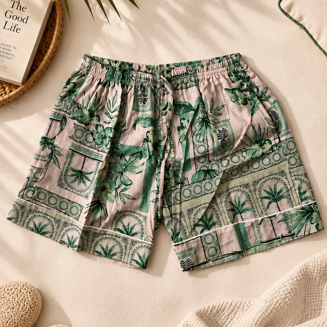 A pair of women's shorts with a green tropical palm tree and geometric pattern on a light pink background, styled in a flat-lay with a woven mat, book, slippers, and a knit blanket.