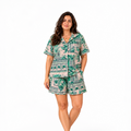A woman modeling a matching short-sleeve button-up pajama shirt and shorts set with a green tropical and architectural scenic print on a light beige background, shot against a solid white studio background.