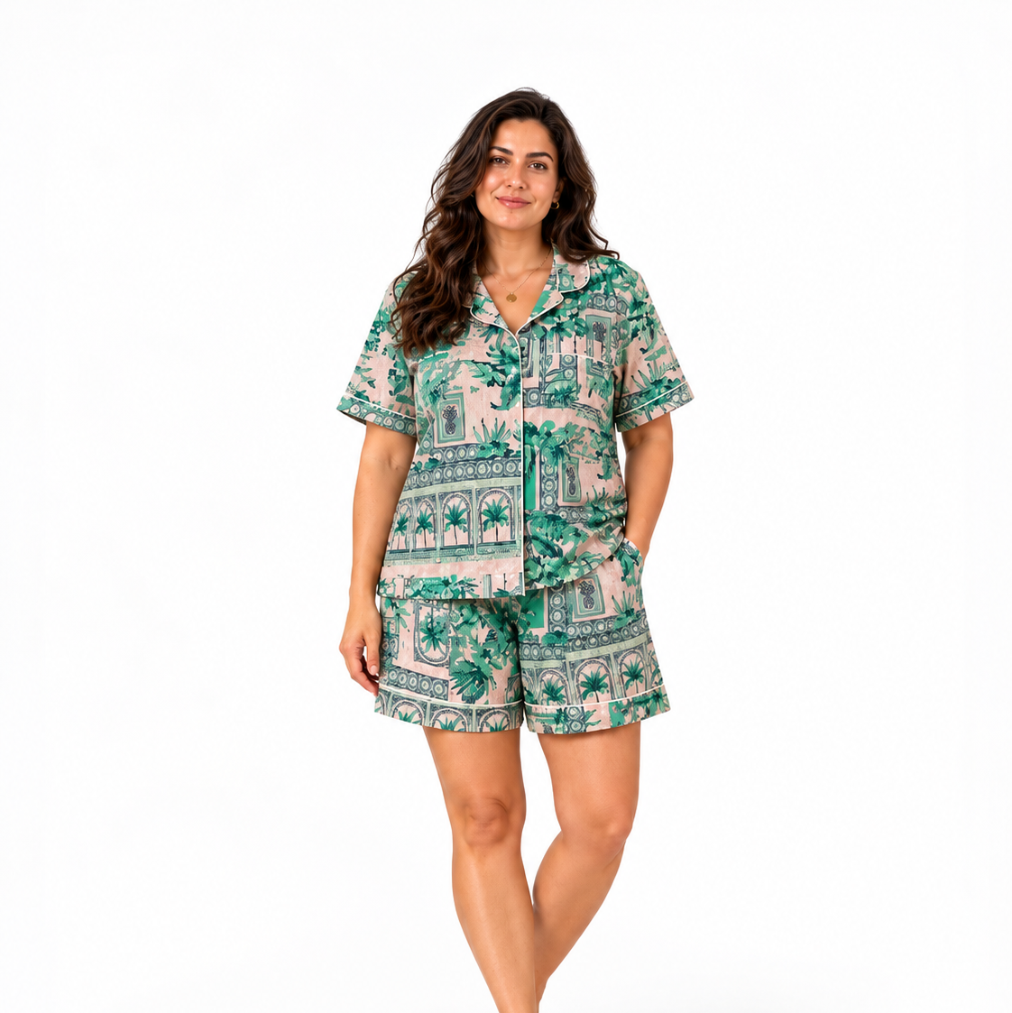 A woman modeling a matching short-sleeve button-up pajama shirt and shorts set with a green tropical and architectural scenic print on a light beige background, shot against a solid white studio background.