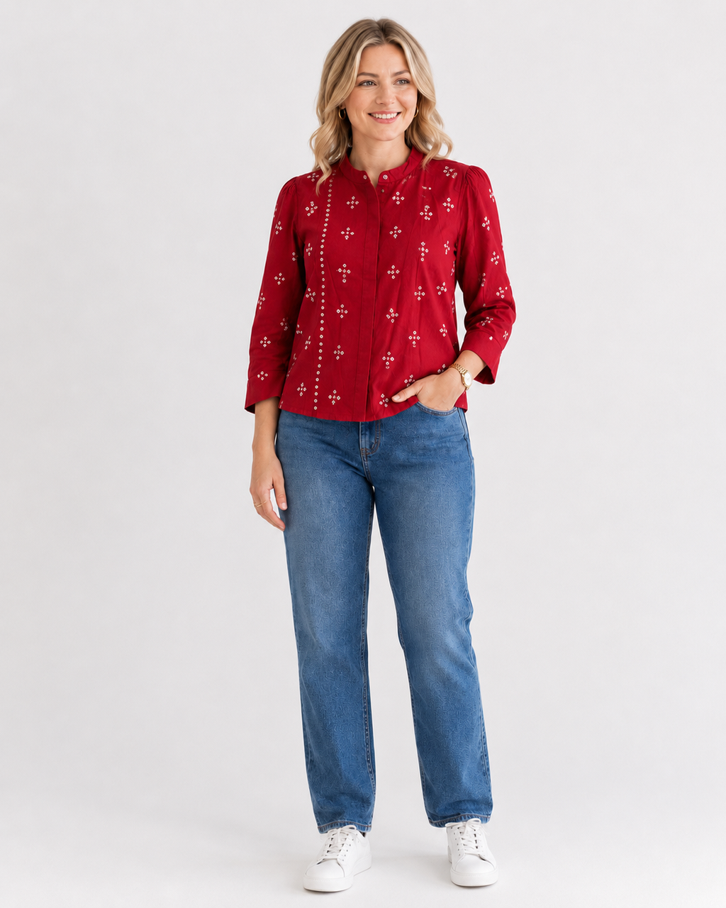 A woman modeling a red long-sleeve button-up shirt with white embroidered cross patterns, paired with blue jeans and white sneakers, against a light gray studio background.