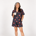 A woman modeling a short-sleeve floral pajama set with a matching top and shorts, holding a white mug against a plain white studio background.