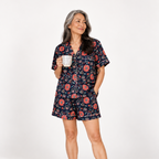 A woman modeling a short-sleeve floral pajama set with a matching top and shorts, holding a white mug against a plain white studio background.