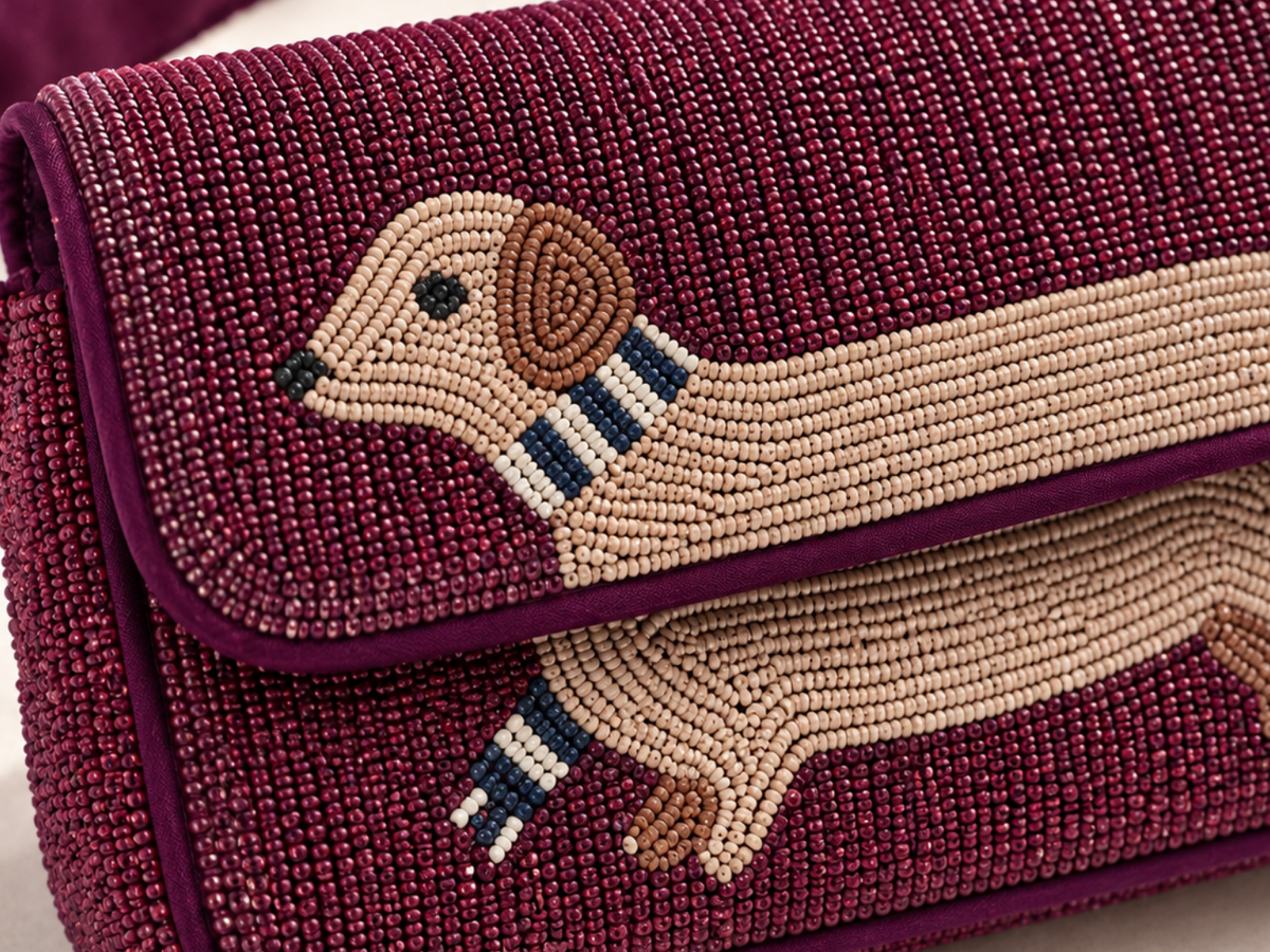 A close-up of a beaded shoulder bag in deep burgundy with a dachshund design on the flap. The bag features intricate beadwork in burgundy, beige, brown, navy, and white, with a matching burgundy strap visible in the upper left corner.