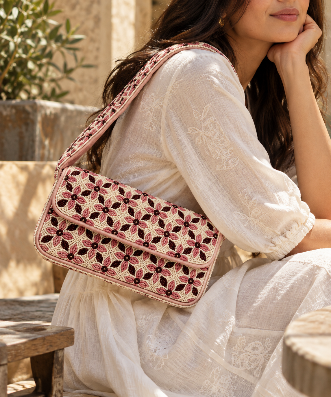 A woman in a white embroidered dress carries a pink and beige embroidered shoulder bag with a floral pattern in an outdoor setting.