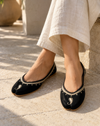 A person wearing black split-toe flat shoes with silver paisley embroidery and a silver beaded border, paired with a cream linen skirt, sitting outdoors on a stone surface.