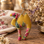 A small fabric camel keychain with a mustard yellow base and a pink, green, and red floral pattern, attached to a silver metal split ring, shown on a plain off-white background.