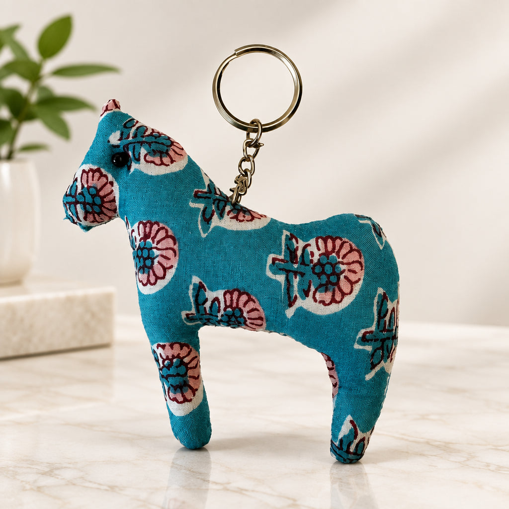 A small fabric horse-shaped keychain charm in teal blue with a pink, white, and blue floral pattern, attached to a silver metal key ring, shown on a plain off-white background.
