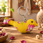 A small bird-shaped fabric keychain charm in yellow with pink lotus flowers, attached to a silver metal key ring, shown on a plain light background.