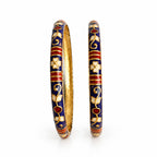 A pair of traditional Indian Kundan bangles with a deep blue enamel outer body, decorated with gold floral vines, red accents, and white enamel flowers, showing the polished gold inner surface.