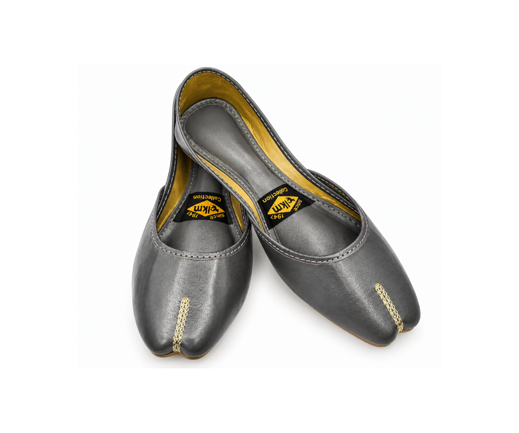 A pair of dark gray leather Juttis with gold-colored decorative stitching on the toes, displayed on a clean white background. The brand label 'KUM' is visible inside the shoes.