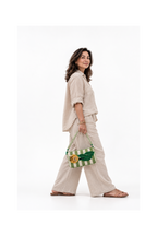 A woman wearing a beige linen button-up shirt and matching wide-leg pants walks while carrying a green and white striped woven handbag with a green leaf patch.