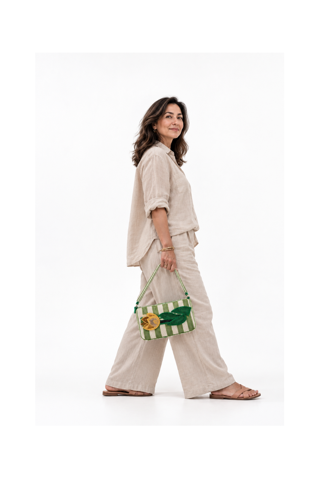 A woman wearing a beige linen button-up shirt and matching wide-leg pants walks while carrying a green and white striped woven handbag with a green leaf patch.