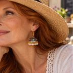 A woman with long red hair wearing a straw sun hat with a black band and ornate gold jhumka earrings, resting her chin on her hand at an outdoor cafe.